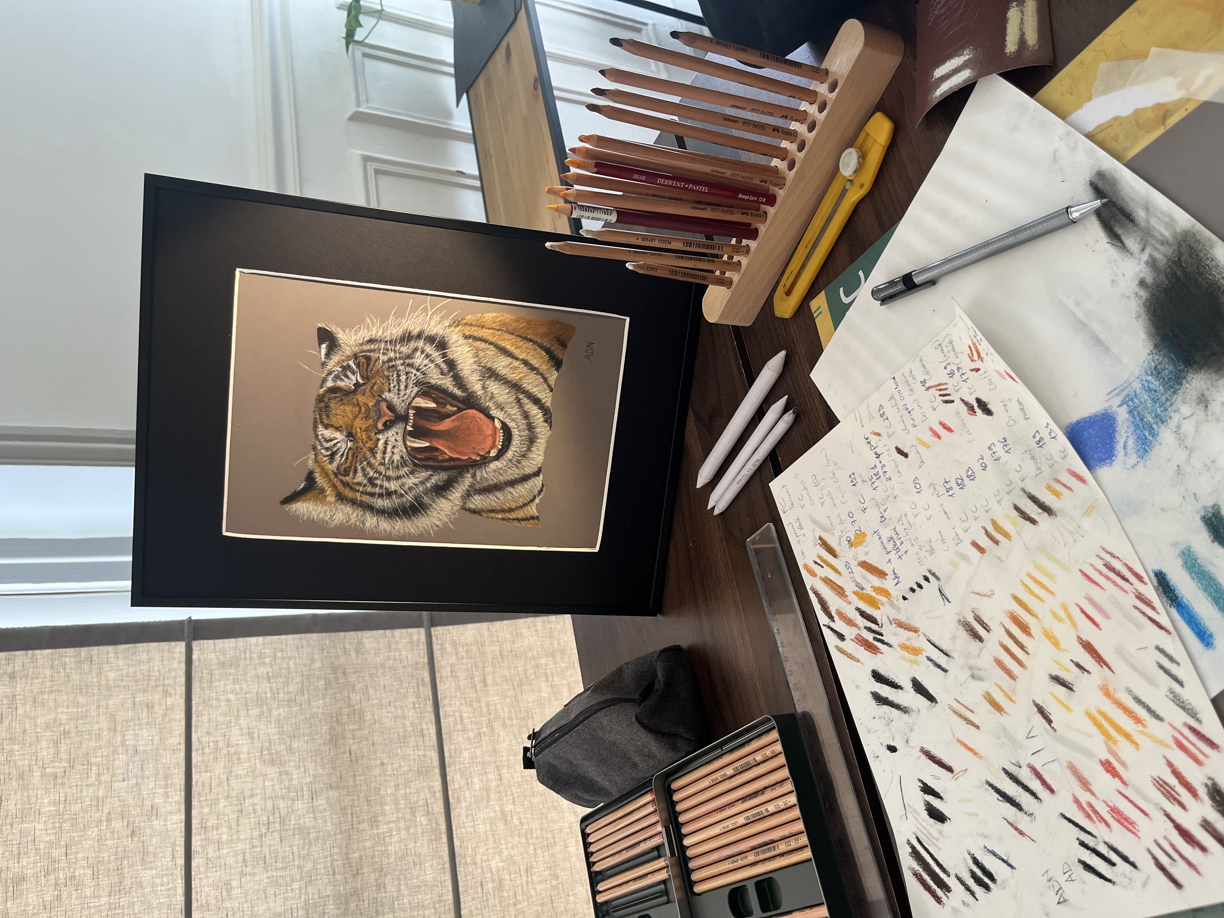 Dessin réaliste au pastel sec d’un tigre rugissant, mis en situation sur un bureau avec du matériel de dessin, collection Félins par ADN - ADN Portfolio "Éclat Sauvage"